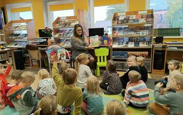 Dzieci siedzą na dywanie i karimatach i patrzą w stronę bibliotekarki, kt&oacute;ra im pokazuje okładkę książki.