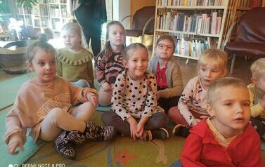 Dzieci siedzą na dywanie i karimatach i oglądają na dużym ekranie film o 1 listopada.