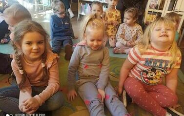 Dzieci siedzą na dywanie i ogłądają na dużym ekranie film o 1 listopada.