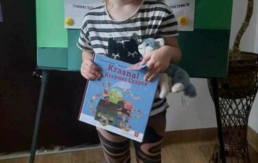 Dziewczynka trzyma w rękach swoją nową książkę z wyprawki czytelniczej z kartą Młodego Czytelnika. Za Nią jest tablica z plakatem promującym akcję MAŁA KSIĄŻKA WIELKI CZŁOWIEK.