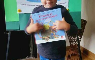 Chlopiec stoi ze swoją książeczką z wyprawki czytelniczej, za nim jest tablica z plakatem promującym akcję MAŁA KSIĄŻKA WIELKI CZłOWIEK.