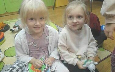 Dwie dziewczynki o blond włosach siedzą na dywanie i trzymają w rękach pluszowe książeczki, w tel widać inne bawiące się dzieci.