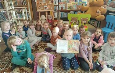 Dzieci siedzą na dywanie, jedna z dziewczynek ma otwarte książkę na bajce o misiach i ją pokazuje.