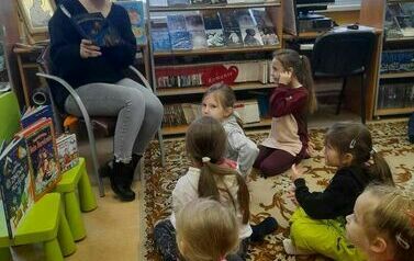 Grupa dzieci siedzących na dywanie i patrzących na bibliotekarkę, kt&oacute;ra czyta im bajkę.