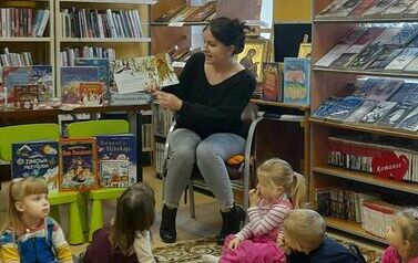 Dzieci siedzą na dywanie odwr&oacute;cone tyłem i patrzą w stronę bibliotekarki, kt&oacute;ra czyta im bajkę.