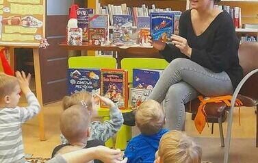 Bibliotekarka siedzi z przodu przy stoliku z książeczkami i pokazuje dzieciom książeczkę o Mikołaju. Niektore dzieci podnoszą ręce w g&oacute;rę chcąc coś powiedzieć.