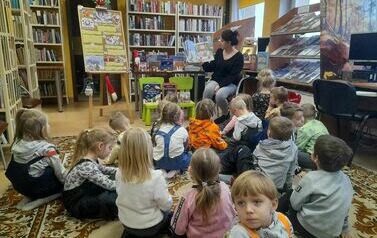 Dzieci siedzą na dywanie odwr&oacute;cone tyłem i patrzą w stronę bibliotekarki, kt&oacute;ra czyta im bajkę.