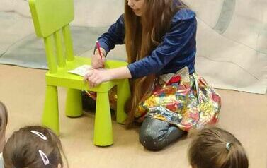 Dzieci patrzą na teatrzyk gdzie Pani przebrana za dziewczynkę pisze coś na kartce na małym zielonym krzesełku.