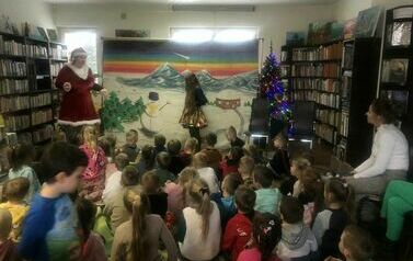 Duża grupa dzieci siedzi na dywanie, z boku siedzi nauczycielka. Na tle zaśnieżonych g&oacute;r widać Mikołaja i dziewczynkę.