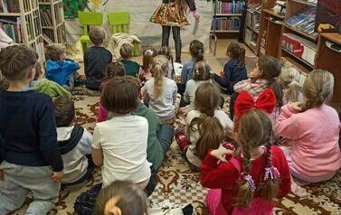 Dzieci siedzą na dywanie i patrzą na teatrzyk. Z przodu stoi aktorka przebrana za dziewczynkę na tle zaśnieżonych g&oacute;r.