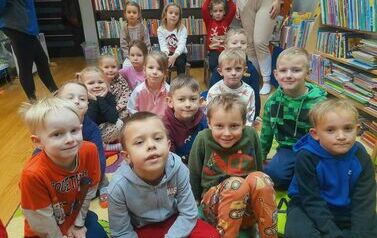 Dzieci siedzą na dywanie i patrzą na bibliotekarkę, kt&oacute;ra im czyta bajkę.