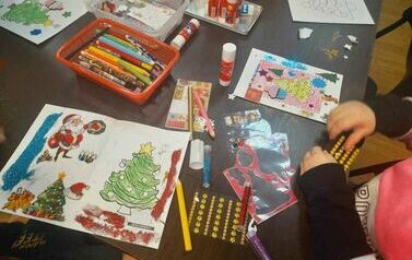 Na stole leżą kredki, kleje, brokat, naklejki i cekinki oraz prace dzieci w trakcie robienia.