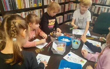 Dzieci siedzą przy stole i robią kartki świąteczne przy pomocy kredek, naklejek, brokatu i cekink&oacute;w.