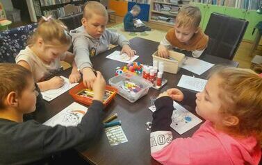 Dzieci siedzą przy stole i robią kartki świąteczne przy pomocy kredek, naklejek, brokatu i cekink&oacute;w.