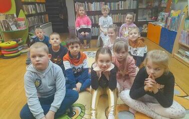 Dzieci siedzą na dywanie i patrzą na bibliotekarkę, kt&oacute;ra im czyta bajkę.