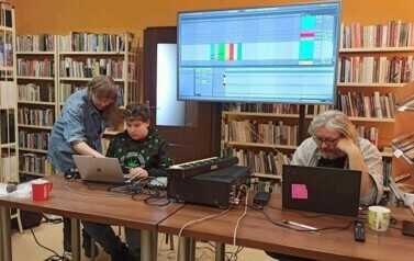 Chłopiec siedzi przy sprzęcie muzycznym, przy nim pochylona jest artystka coś mu pokazując. Z prawej strony siedzi muzyk patrząc w laptopa.