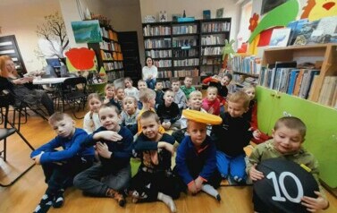 Cała grupa dzieci siedzi na dywanie i słucha bajki czytanej przez bibliotekarkę. Za nimi na krzesełku siedzi nauczycielka.