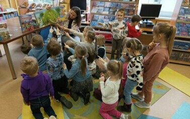 Grupa dzieci, kt&oacute;ra ma skierowane rączki do książki w 3D o dinozaurach przy kt&oacute;rej siedzi bibliotekarka pokazując wnętrze książki. 