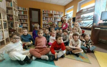 Zdjęcie grupowe dzieci z bibliotekarką z tyłu, kt&oacute;ra trzyma dziewczynkę na kolanie i w ręku książeczkę o dinozaurach.