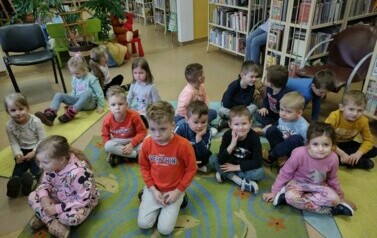 Dzieci siedzą na dywanie i karimatach i słuchają bajki, kt&oacute;rą czyta im bibliotekarka.