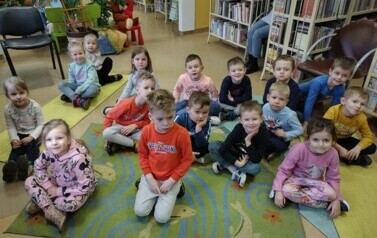 Dzieci siedzą na dywanie i karimatach i słuchają bajki, kt&oacute;rą czyta im bibliotekarka.