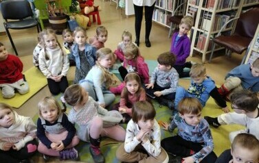 Dzieci siedzą na dywanie i karimatach i słuchają bajki, kt&oacute;rą czyta im bibliotekarka.
