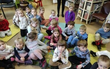 Dzieci siedzą na dywanie i karimatach i słuchają bajki, kt&oacute;rą czyta im bibliotekarka.
