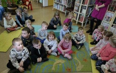 Dzieci siedzą na dywanie i karimatach i słuchają bajki, kt&oacute;rą czyta im bibliotekarka.