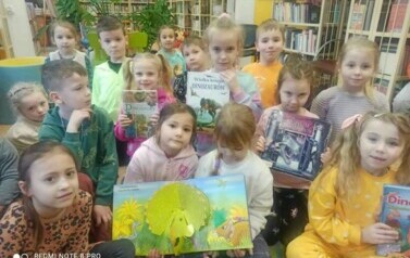 Dzieci siedzą w grupie, niekt&oacute;re z nich trzymają książeczki pokazując je przodem, na środku dwie dziewczynki mają otwarte książkę o dinozaurach w formacie 3D.
