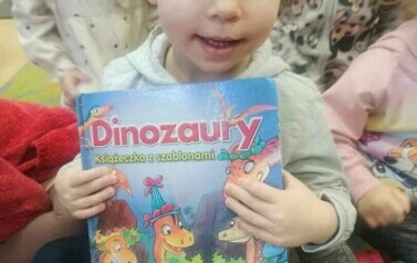 Dzieci siedzą na dywanie i słuchają bajki. Z przodu siedzi dziewczynka trzymając w rączkach książkę o dinozaurach pokazując ją przodem do zdjęcia.