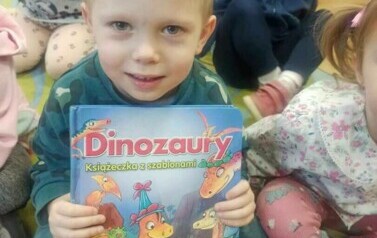Dzieci siedzą na dywanie i słuchają bajki. Z przodu siedzi chłopiec trzymając w rączkach książkę o dinozaurach pokazując ją przodem do zdjęcia.
