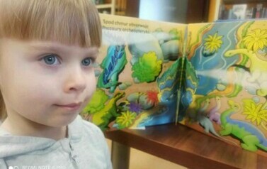 Dziewczynka w blond włosach stoi obok otwartej książeczki w kt&oacute;rej widać dinozaury w formacie 3D.