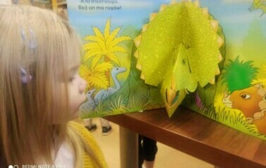 Dziewczynka w blond włosach patrzy w książeczkę w kt&oacute;rej widać dinozaura w formacie 3D.