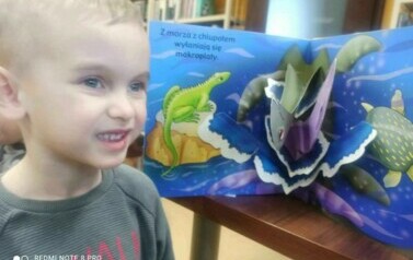 Chłopiec w blond włosach stoi obok otwartej książeczki  w kt&oacute;rej widać dinozaura w formacie 3D.
