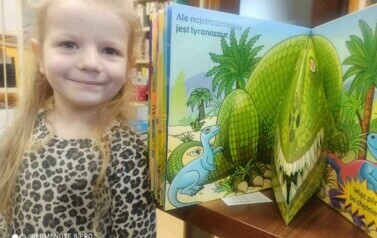 Dziewczynka w blond włosach stoi obok otwartej książeczki w kt&oacute;rej widać dinozaura w 3D.
