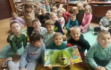 Grupa dzieci siedzi na karimatach, z przodu po środku chłopiec trzyma otwartą książkę o dinozaurach w 3D.