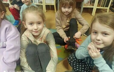 Z przodu na dywanie siedzą trzy dziewczynki słuchające bajki. W tle widać inne dzieci.