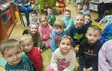 Grupa dzieci siedzi na dywanie i słucha bajki, kt&oacute;rą czyta bibliotekarka.