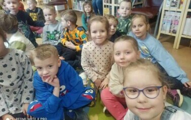 Dzieci siedzą na dywanie i słuchają bajki czytanej przez bibliotekarkę.