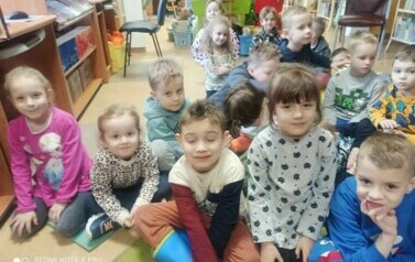 Grupa dzieci siedzi na dywanie i słucha bajki, kt&oacute;rą czyta bibliotekarka.