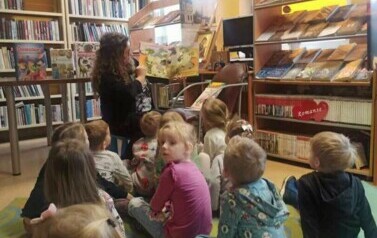 Dzieci siedza na dywanie i patrzą w stonę bibliotekarki, kt&oacute;ra czyta i pokazuje stronice bajki o dinozaurach.