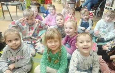 Grupa dzieci siedzi na dywanie i słucha bajki, kt&oacute;rą czyta bibliotekarka.