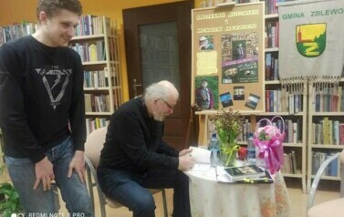Pisarz przy stoliku podpisuje dedykację dla czytelnika, kt&oacute;ry stoi obok i się lekko uśmiecha.