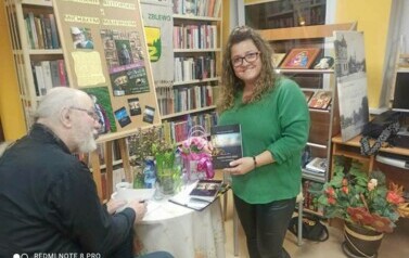 Pisarz przy stoliku podpisuje dedykację na swojej książce dla kierowniczki biblioteki, kt&oacute;ra stoi obok i się uśmiecha trzymając w rękach jedną z książek autora.