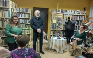 Z przodu stoją : pisarz, kierownik biblioteki i fotograf. Przy stoliku siedzi prowadząca. Widać kilka czytelnik&oacute;w.