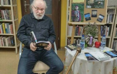 Pisarz siedzi przy stoliku i czyta fragment swojej książki. O stolik jest oparta Jego laska.