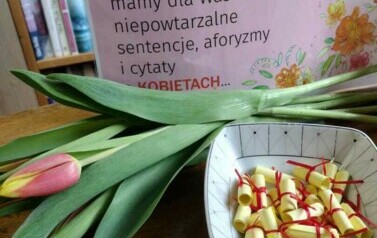 Na stole leżą tulipany, aforyzmy zwinięte w rulonik i zalaminowana kartka z cytatem o kobietach.