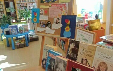 Na biurku są wyłożone książki o kobietach, cukierki i aforyzmy zwinięte w rulonik. Obok biurka stoi przystrojona tablica korkowa w plakat i ciekawostki o Dniu Kobiet.