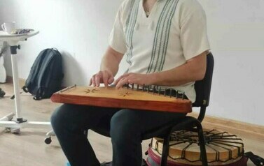 Szymon siedzi na krześle i gra na jakimś starym drewnianym instrumencie.