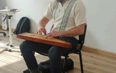 Szymon siedzi na krześle i gra na jakimś starym drewnianym instrumencie.